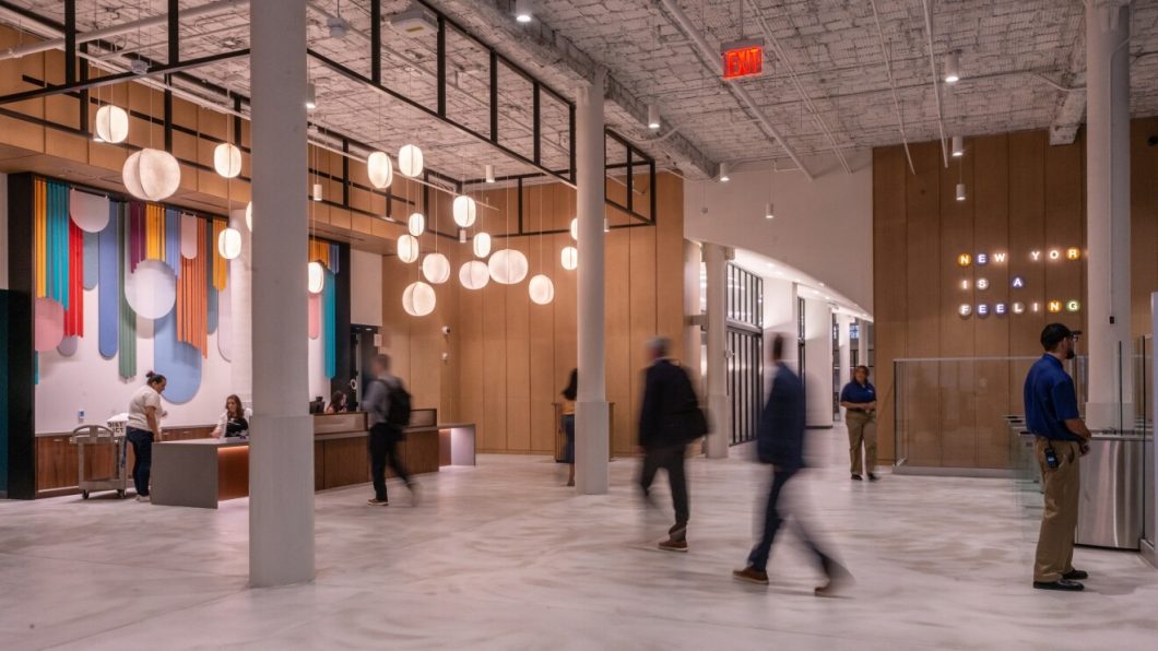 Fotografia de um amplo saguão moderno e iluminado. No lado esquerdo, um balcão de recepção de madeira com funcionários e pessoas em movimento. Atrás do balcão, uma parede decorada com formas geométricas coloridas em tons de azul, rosa, amarelo e roxo. Pendentes esféricos brancos descem do teto industrial de tijolos brancos. À direita, uma parede de madeira exibe o letreiro luminoso colorido "NEW YORK IS A FEELING". Pessoas borradas caminham pelo piso de concreto claro polido entre colunas brancas.