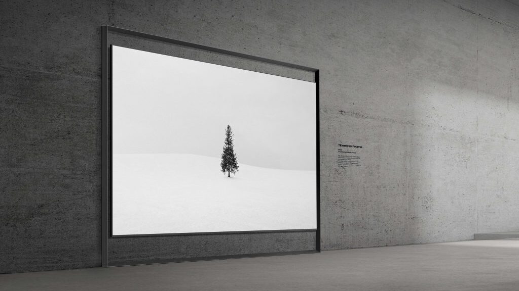 Painel de exibi&ccedil;&atilde;o digital de arte instalado rente a uma parede de concreto bruto em uma galeria ou ambiente minimalista. A tela, emoldurada por uma estrutura fina e elegante, mostra uma imagem minimalista de uma &aacute;rvore isolada no gelo. Ao lado da tela, h&aacute; uma pequena inscri&ccedil;&atilde;o na parede intitulada "Timeless Frame", refor&ccedil;ando o conceito de porta-retratos digital de alta tecnologia.