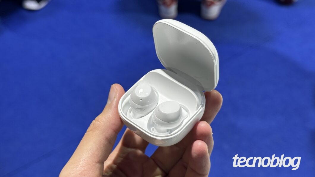 Uma mão segura um estojo de fones de ouvido sem fio de cor branca, que está aberto e revela dois fones também brancos. O logotipo "tecnoblog" está no canto inferior direito, em branco.