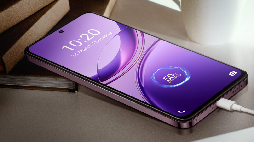 Smartphone de bordas retas em tom roxo met&aacute;lico apoiado sobre uma mesa branca, conectado a um cabo de carregamento branco. Na tela, com fundo roxo e gr&aacute;fico circular azul, aparece o hor&aacute;rio "10:20" e a data "24 March Thursday", al&eacute;m do indicador de bateria com "50% charging". Ao fundo, h&aacute; dois cadernos marrons empilhados e uma caneca branca vazia. A ilumina&ccedil;&atilde;o &eacute; suave, vinda da direita da imagem.