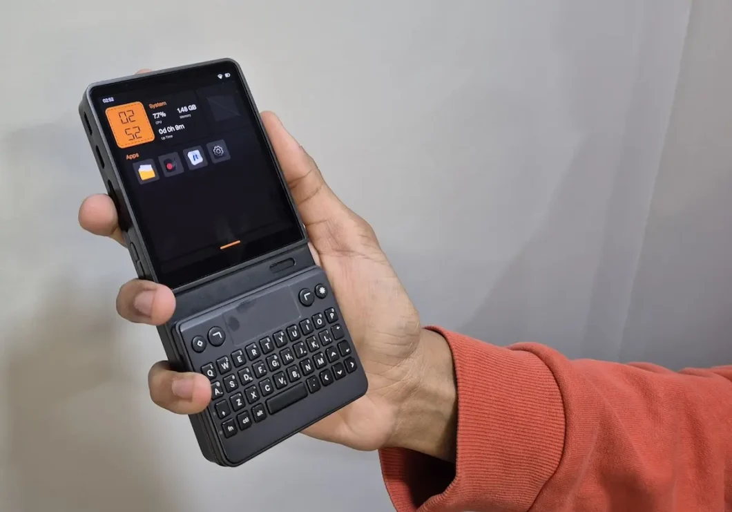 Uma m&atilde;o segura o computador de bolso Mecha Comet em frente a uma parede cinza. O aparelho &eacute; preto e possui uma tela sens&iacute;vel ao toque com interface escura e detalhes laranjas. Abaixo do visor, h&aacute; um m&oacute;dulo de teclado f&iacute;sico QWERTY preto com bot&otilde;es de navega&ccedil;&atilde;o. Na lateral esquerda, s&atilde;o vis&iacute;veis bot&otilde;es de comando. A pessoa veste um casaco de cor terracota. A imagem destaca a portabilidade do dispositivo e seu formato vertical.