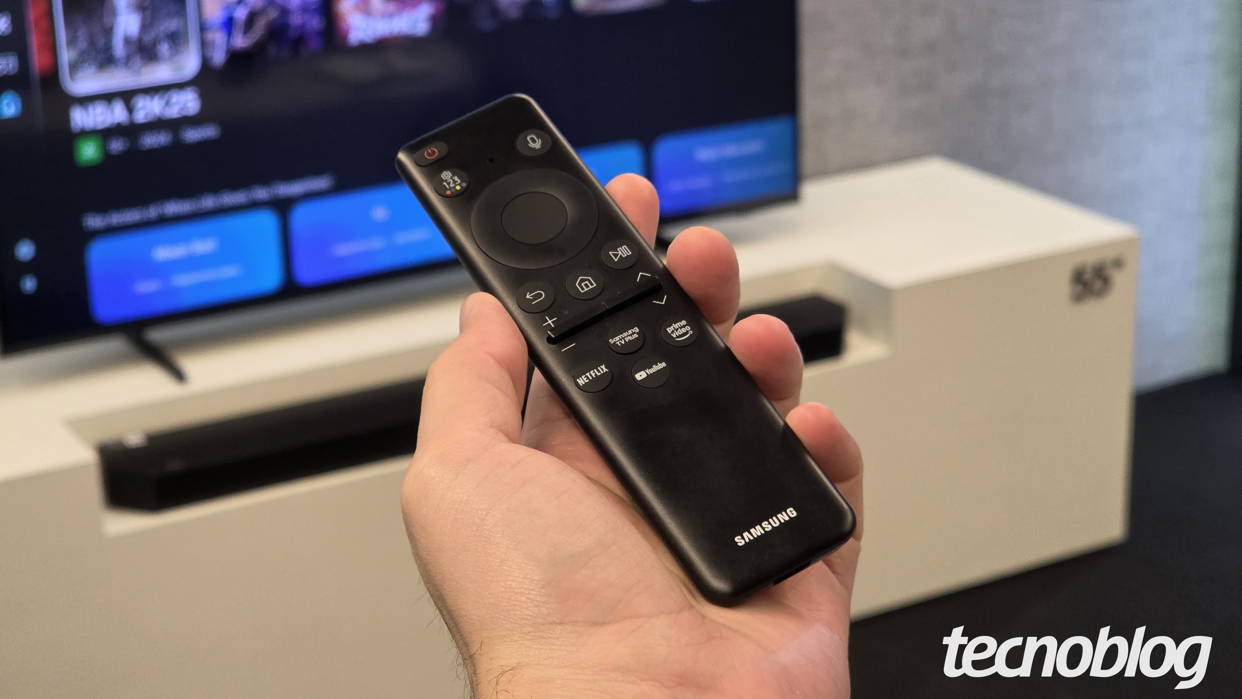 M&atilde;o segura controle remoto preto Samsung com bot&otilde;es de voz e acesso r&aacute;pido a Netflix, YouTube e Prime Video, TV ao fundo.