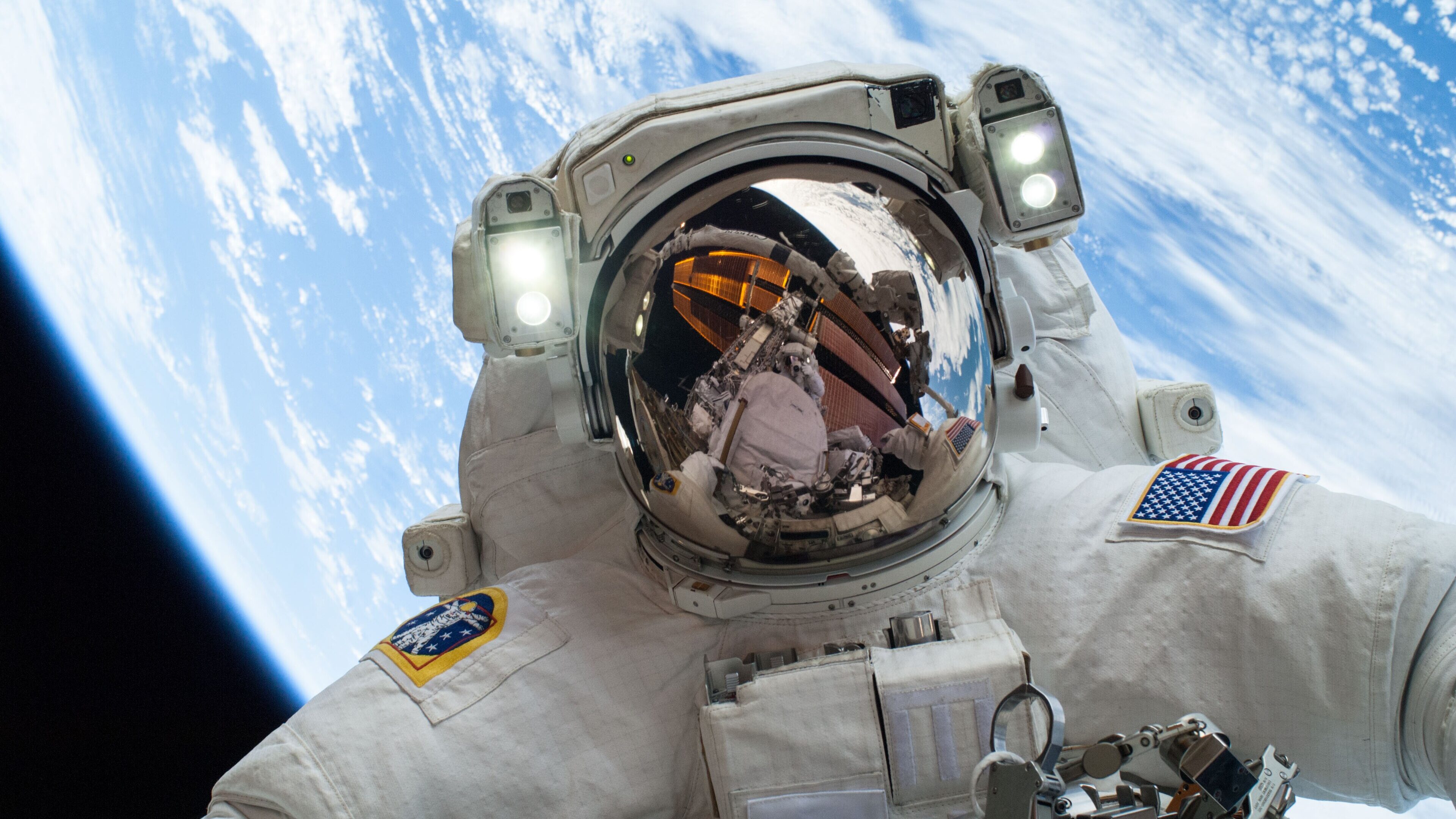 Selfie em close de um astronauta em traje espacial branco durante caminhada espacial. O visor do capacete reflete a estrutura da esta&ccedil;&atilde;o e parte do planeta Terra. Duas luzes brancas intensas est&atilde;o acesas acima do visor. No bra&ccedil;o esquerdo, h&aacute; uma bandeira dos Estados Unidos e, no direito, um emblema circular colorido. O fundo exibe a curvatura da Terra com tons de azul intenso e nuvens brancas sobre o espa&ccedil;o negro. Na parte inferior, veem-se ferramentas e partes met&aacute;licas do traje.