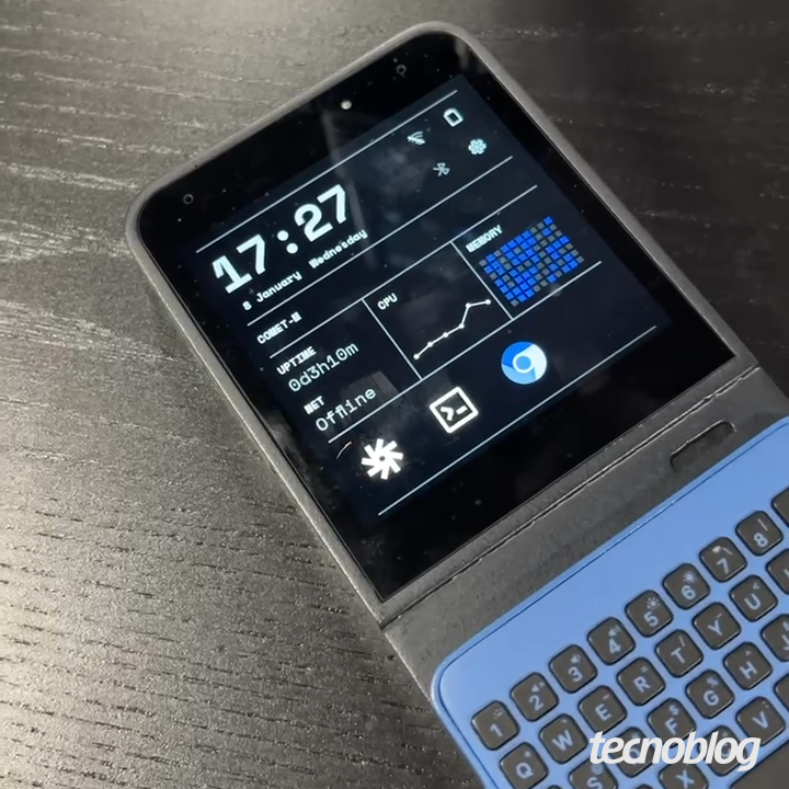 Dispositivo retangular preto sobre mesa escura. A tela exibe um painel de sistema com o hor&aacute;rio "17:27" e widgets de desempenho. Na metade inferior, est&aacute; encaixado um teclado f&iacute;sico modular na cor azul-claro com teclas cinzas. O design &eacute; compacto, assemelhando-se a um smartphone espesso com acess&oacute;rios acoplados.