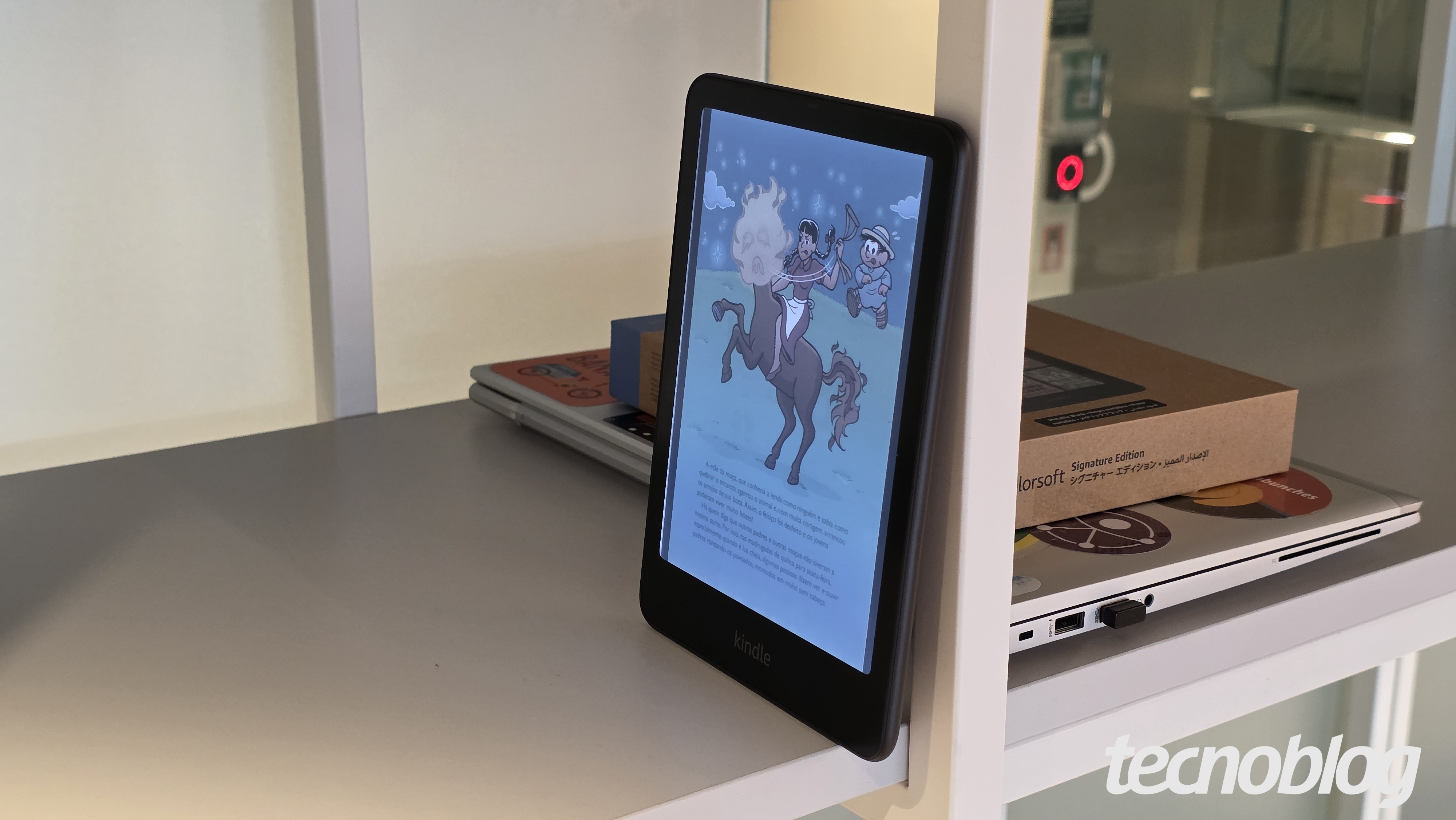 Kindle Colorsoft apoiado em prateleira, com notebook e a caixa do Kindle Colorwhite atr&aacute;s