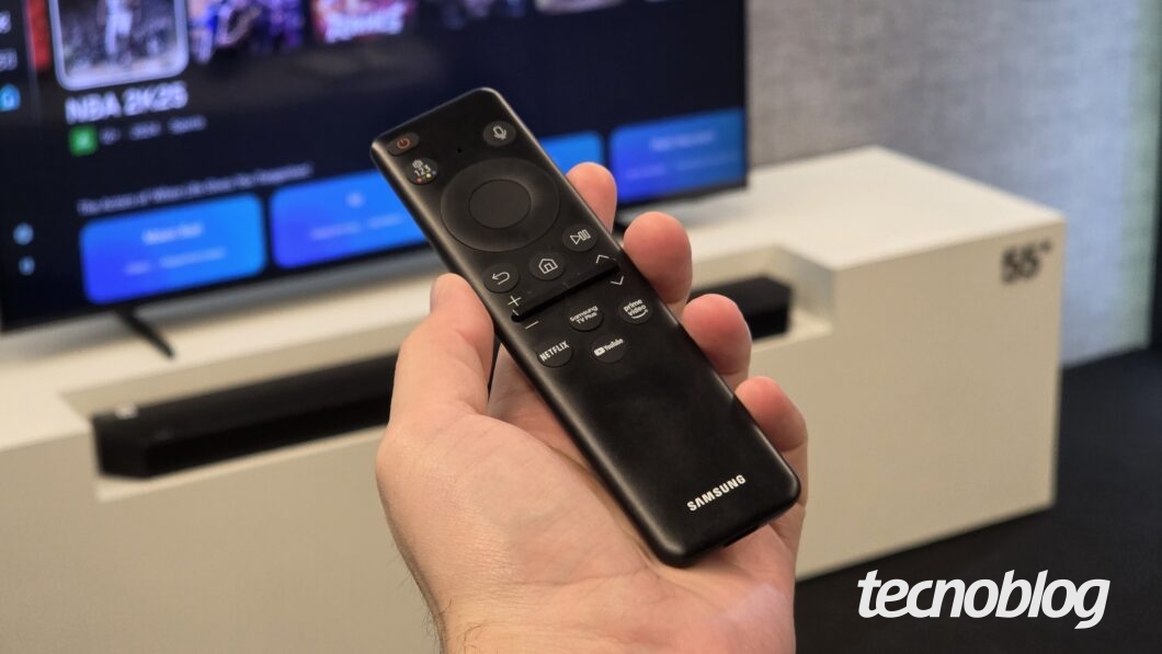 M&atilde;o segura controle remoto preto Samsung com bot&otilde;es de voz e acesso r&aacute;pido a Netflix, YouTube e Prime Video, TV ao fundo.