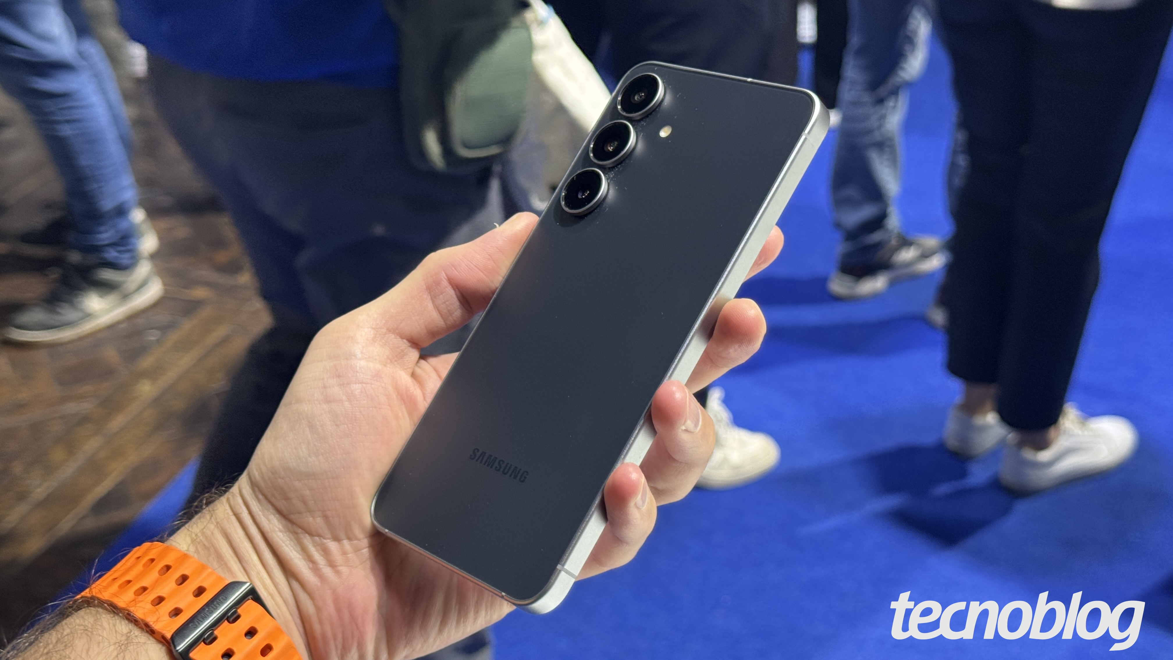 Uma m&atilde;o segura um smartphone Galaxy S25 FE preto e prata, exibindo a parte de tr&aacute;s do aparelho. O telefone tem tr&ecirc;s c&acirc;meras alinhadas verticalmente no canto superior esquerdo e o logotipo "SAMSUNG" na parte inferior. A m&atilde;o tem uma pulseira de rel&oacute;gio laranja. O fundo &eacute; desfocado, mostrando pessoas e um piso azul. Na parte inferior direita, a marca d'&aacute;gua do "tecnoblog" &eacute; vis&iacute;vel.