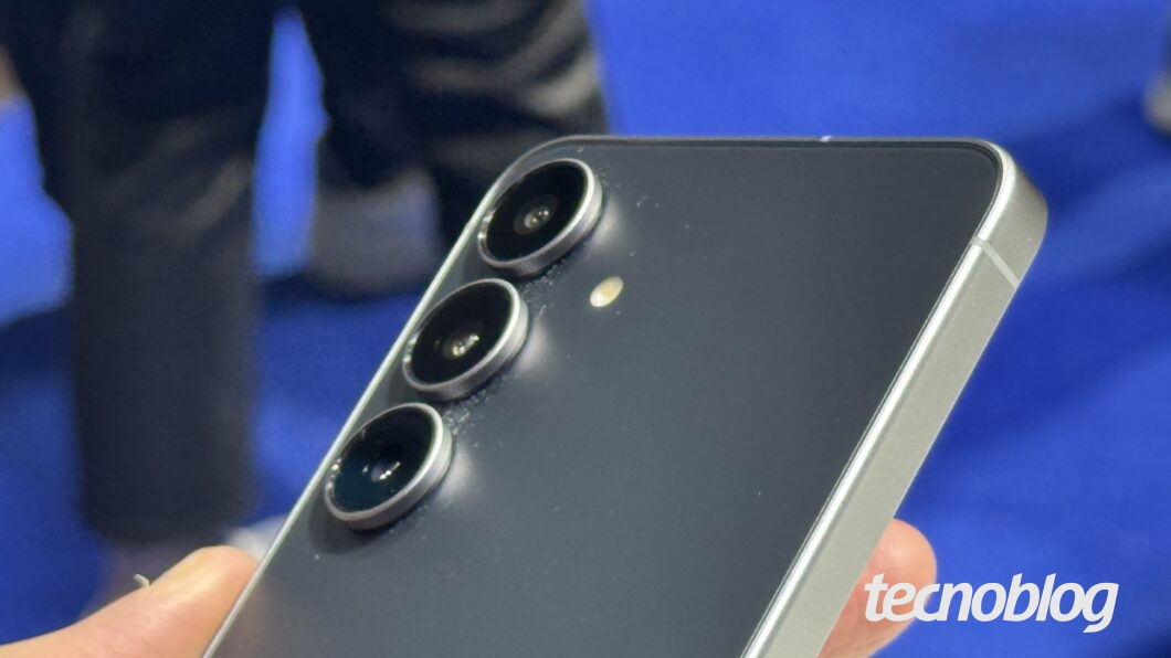 Uma m&atilde;o segura um smartphone Galaxy S25 FE cinza e preto. O aparelho &eacute; exibido de perfil, mostrando o acabamento met&aacute;lico e o m&oacute;dulo de c&acirc;mera com tr&ecirc;s lentes circulares alinhadas verticalmente no canto superior. O flash est&aacute; localizado ao lado das lentes. O fundo &eacute; desfocado, mostrando pessoas e um piso azul. No canto inferior direito, o logotipo "tecnoblog" est&aacute; em branco.