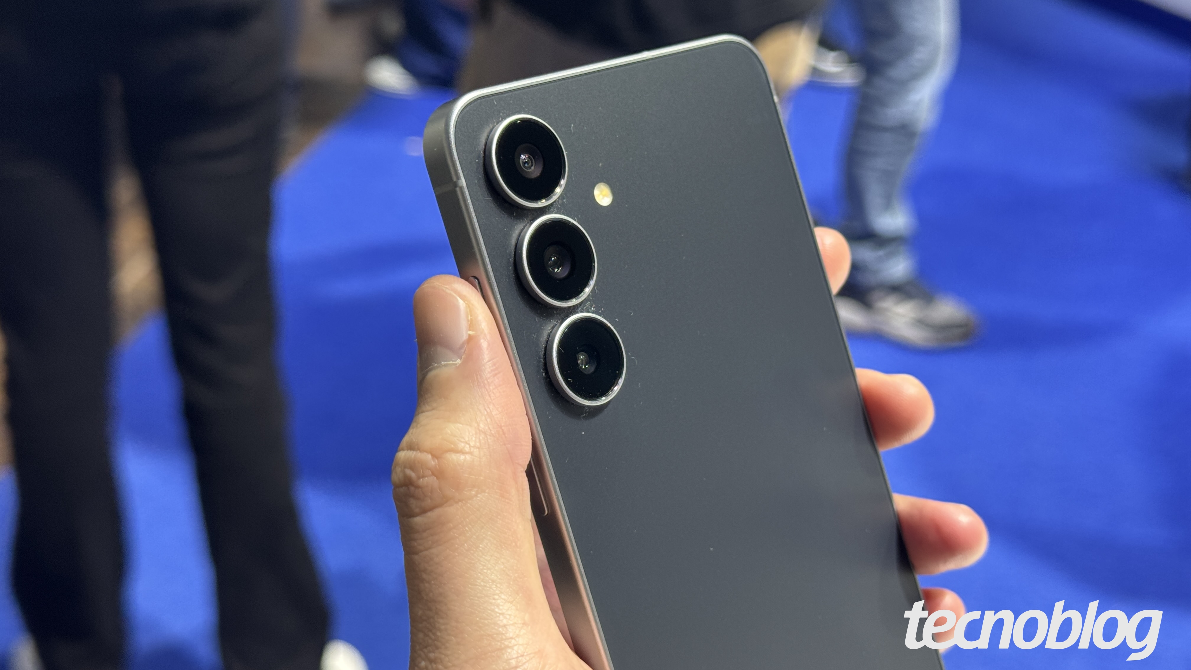 Uma m&atilde;o segura um smartphone Galaxy S25 FE cinza e preto. O aparelho &eacute; exibido de perfil, mostrando o acabamento met&aacute;lico e o m&oacute;dulo de c&acirc;mera com tr&ecirc;s lentes circulares alinhadas verticalmente no canto superior. O flash est&aacute; localizado ao lado das lentes. O fundo &eacute; desfocado, mostrando pessoas e um piso azul. No canto inferior direito, o logotipo "tecnoblog" est&aacute; em branco.