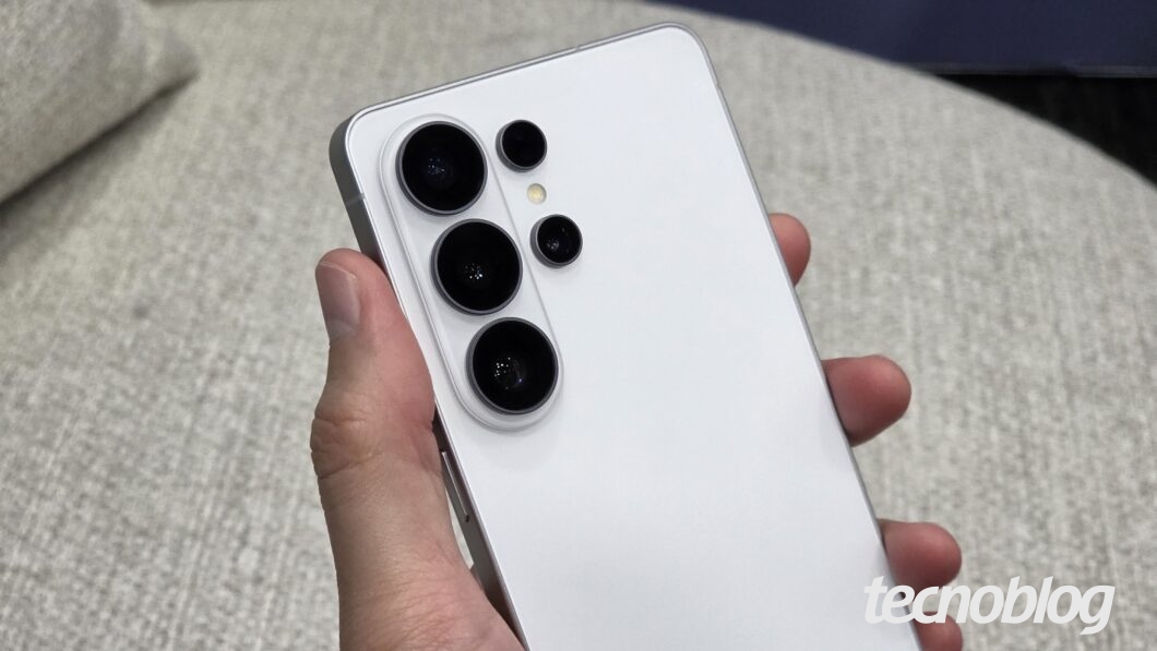 Close-up da parte traseira de um smartphone na cor cinza-claro, segurado por uma mão. O destaque são as cinco aberturas circulares para câmeras e sensores, alinhadas verticalmente em duas colunas: três lentes maiores à esquerda e duas menores à direita, com um flash LED entre elas. O acabamento do aparelho é fosco e as bordas são metálicas e retas. O fundo mostra um estofado cinza desfocado. A marca d'água "tecnoblog" está posicionada de forma semitransparente no canto inferior direito.