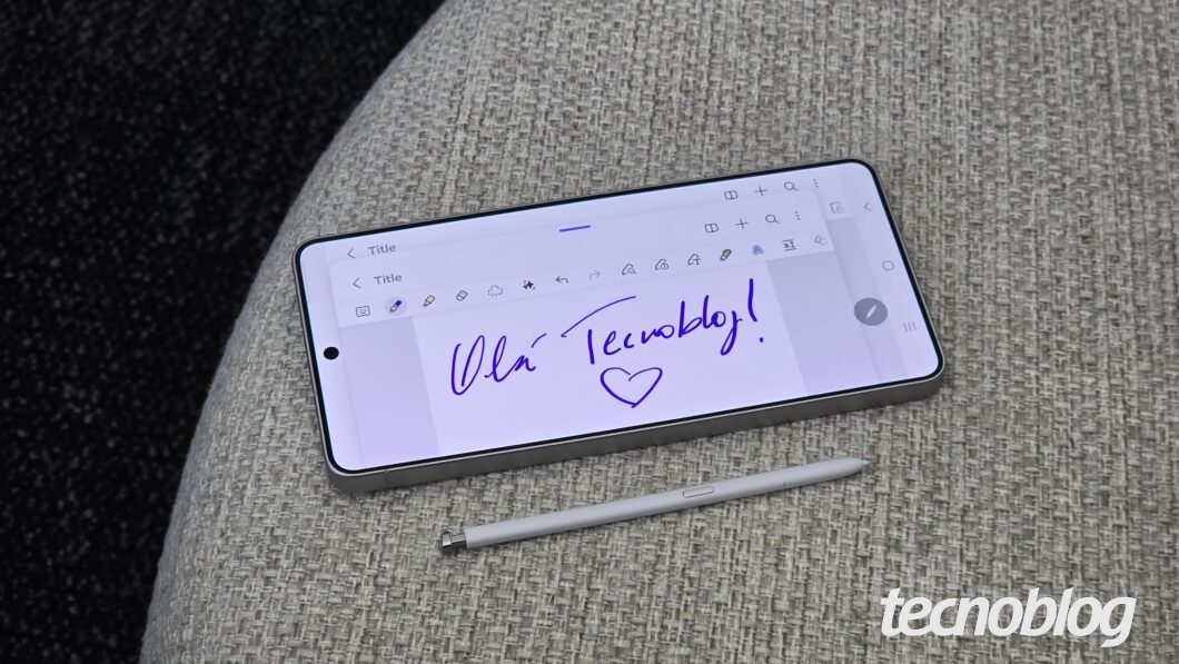Smartphone cinza posicionado horizontalmente sobre um tecido tramado cinza. A tela exibe um aplicativo de notas com fundo branco e a frase manuscrita em azul "Olá Tecnoblog!", acompanhada de um desenho de coração. Abaixo do aparelho, há uma caneta stylus (S Pen) na cor branca com ponta fina e um detalhe metálico no topo. A interface da tela mostra ícones de ferramentas de edição e botões virtuais de navegação. No canto inferior direito da imagem, consta a marca d'água "tecnoblog".