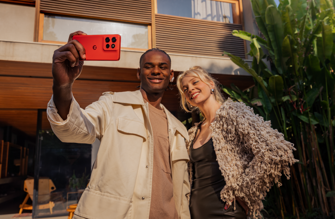 Duas pessoas posam para uma selfie ao ar livre enquanto uma delas segura um smartphone Motorola vermelho, voltado para a c&acirc;mera. O aparelho exibe tr&ecirc;s lentes traseiras e o logotipo da marca. A dupla sorri em frente a uma casa moderna, com plantas tropicais ao fundo e luz natural destacando o design vibrante do celular.
