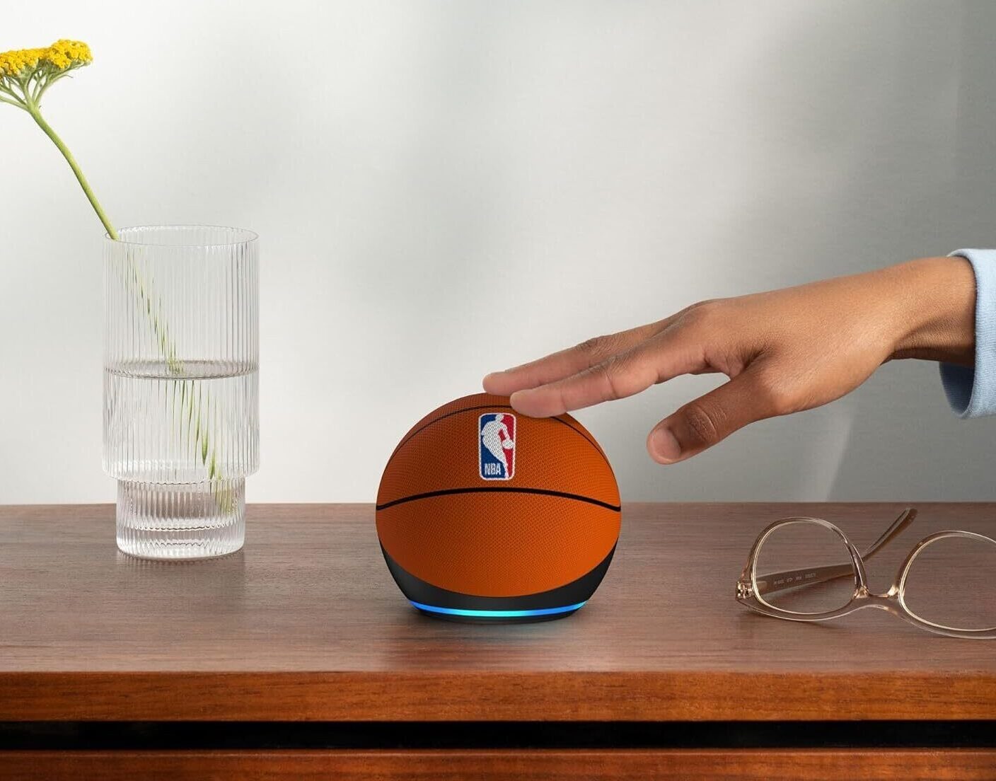 Echo Dot (5&ordf; Gera&ccedil;&atilde;o) em edi&ccedil;&atilde;o limitada NBA, com formato esf&eacute;rico revestido em tecido laranja que imita a textura e as linhas pretas de uma bola de basquete. Na parte superior, destaca-se o logotipo oficial da "NBA" em azul, branco e vermelho. O dispositivo repousa sobre uma mesa de madeira escura. Uma m&atilde;o de pele retinta toca o topo do aparelho. Ao lado, h&aacute; um par de &oacute;culos de arma&ccedil;&atilde;o fina e um vaso de vidro canelado com flores amarelas, contra um fundo de parede cinza claro.