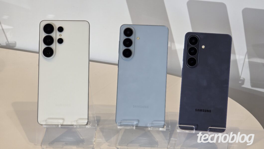 Galaxy S26 Ultra, S26 Plus e S26 vistos de costas. Os aparelhos est&atilde;o sobre suportes transparentes. O S26 Ultra &eacute; o maior deles e tem cor dourada. O S26 Plus &eacute; o intermedi&aacute;rio, um pouco menor que o Ultra, e est&aacute; na cor cinza. O S26 &eacute; o menor dos tr&ecirc;s e est&aacute; na cor preta.