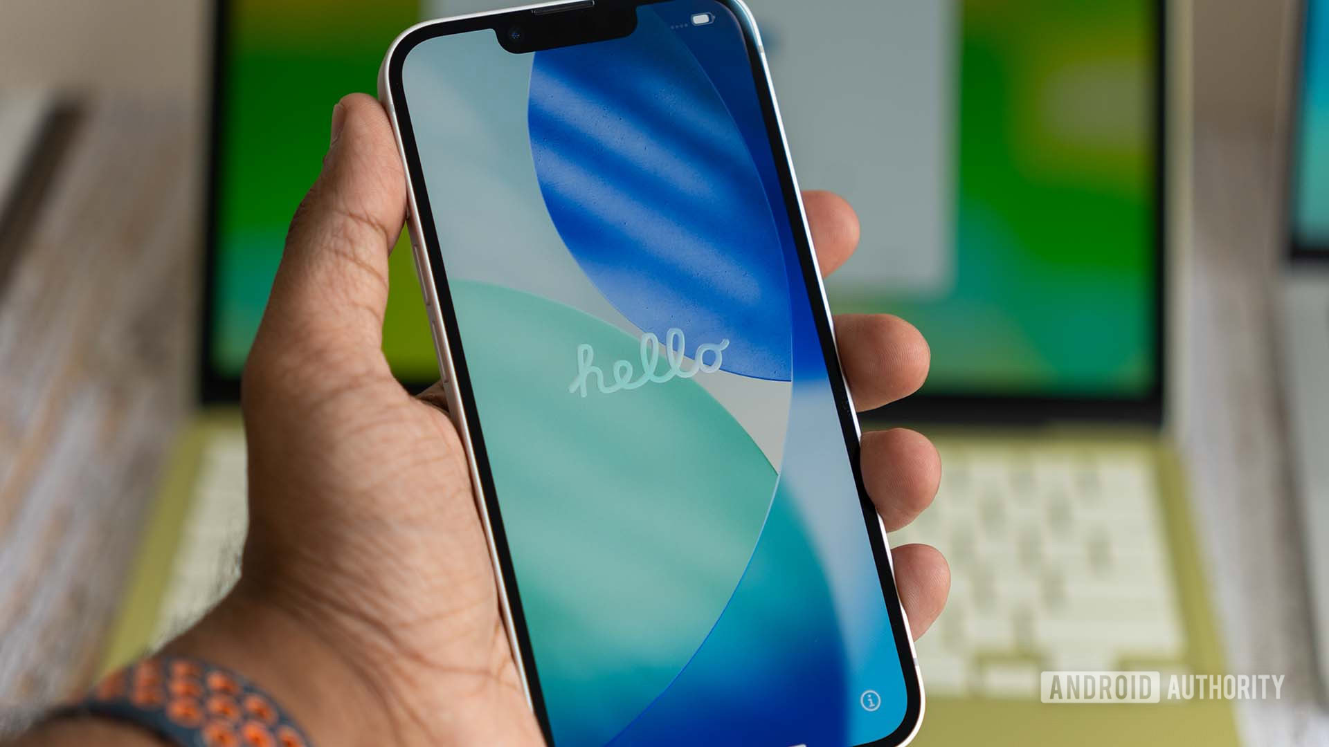 Apple iPhone 17e mostrando a tela de inicializa&ccedil;&atilde;o de Ol&aacute; durante a inicializa&ccedil;&atilde;o, com o MacBook Neo em segundo plano