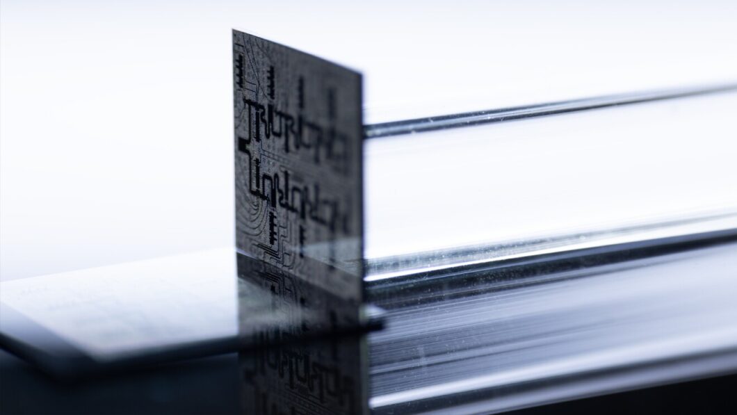 Um chip posicionado verticalmente sobre uma superf&iacute;cie refletiva. O chip &eacute; fino e transl&uacute;cido, com circuitos intricados vis&iacute;veis. A imagem tem um fundo branco e ilumina&ccedil;&atilde;o suave, destacando os detalhes do componente tecnol&oacute;gico.