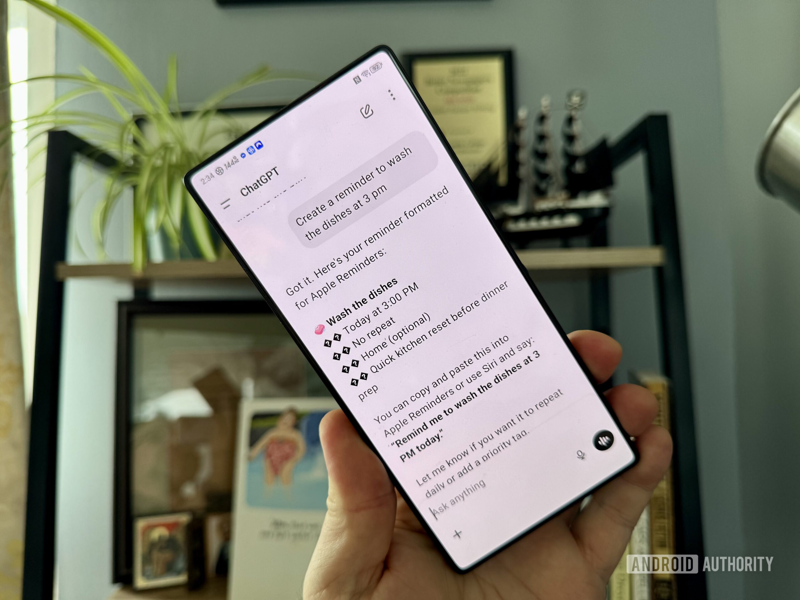 ChatGPT em um telefone Android suspenso no ar pela mão de um homem com uma estante de livros ao fundo.