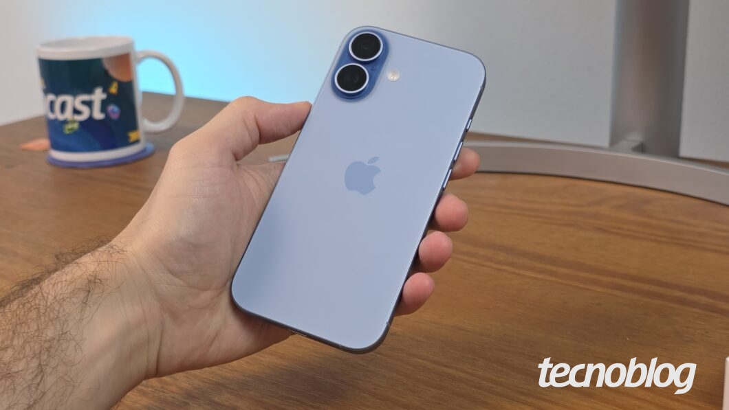 Mão segurando iPhone 17 azul-névoa, com mesa em madeira como fundo