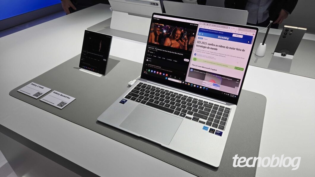 Notebook branco Galaxy Book 5 Pro está sobre uma mesa de exposição branca durante a CES 2025. A tela exibe ícones coloridos, com um show em reprodução numa metade e, na outra, o site do Tecnoblog.