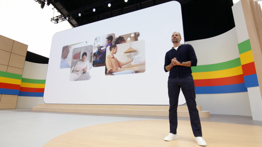 Um homem está de pé em um palco, à direita da imagem. Ele veste um casaco azul-marinho com capuz, calça jeans escura e tênis brancos. Ao fundo, um telão gigante exibe fotos de pessoas utilizando tablets em ambientes domésticos. As paredes laterais do palco são decoradas com faixas horizontais nas cores verde, amarelo, vermelho e azul. O ambiente é amplo, com iluminação de evento tecnológico e teto com estruturas de refletores aparentes.