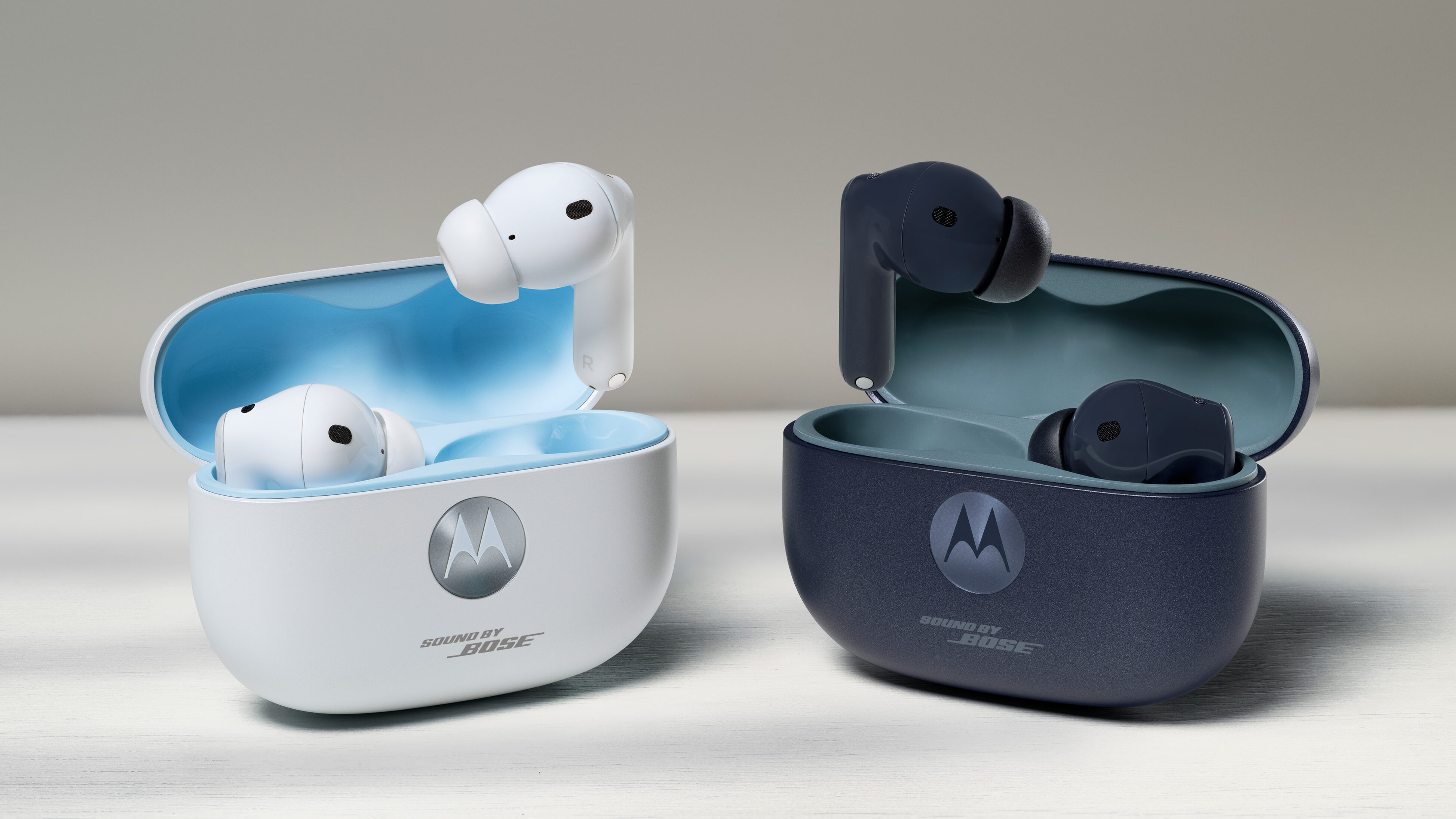 Dois estojos de carregamento de fones de ouvido sem fio da marca Motorola estão dispostos lado a lado sobre uma superfície clara e texturizada. À esquerda, o conjunto é branco com o interior azul-celeste; à direita, é azul-escuro fosco com interior em tom de azul-petróleo. Ambos os estojos estão abertos, revelando um fone encaixado e outro apoiado na borda superior. Na frente de cada estojo, há o logotipo circular da Motorola e a inscrição "SOUND BY BOSE" logo abaixo. Os fones possuem design intra-auricular com hastes curtas.