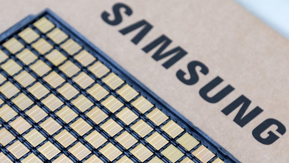 Fotografia em close-up de uma bandeja preta contendo dezenas de pequenos chips semicondutores quadrados dispostos em fileiras organizadas. Cada componente possui uma superfície brilhante com tons de dourado e cobre. Ao lado da bandeja, sobre uma superfície bege, está estampada a palavra "SAMSUNG" em letras maiúsculas pretas e fonte robusta. O foco da imagem está nos chips em primeiro plano, enquanto o nome da marca ao fundo apresenta um leve desfoque, criando profundidade de campo.