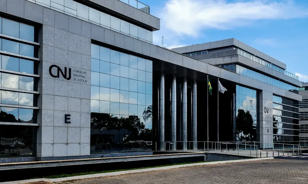Fachada do edifício do Conselho Nacional de Justiça (CNJ), com estrutura de concreto e vidro espelhado. À esquerda, está visível o letreiro com o texto "CNJ Conselho Nacional de Justiça" e a letra "E" abaixo. Ao centro, há colunas metálicas verticais e, à frente delas, duas bandeiras hasteadas: a do Brasil e uma bandeira branca. O céu está parcialmente nublado e se reflete nas janelas espelhadas.