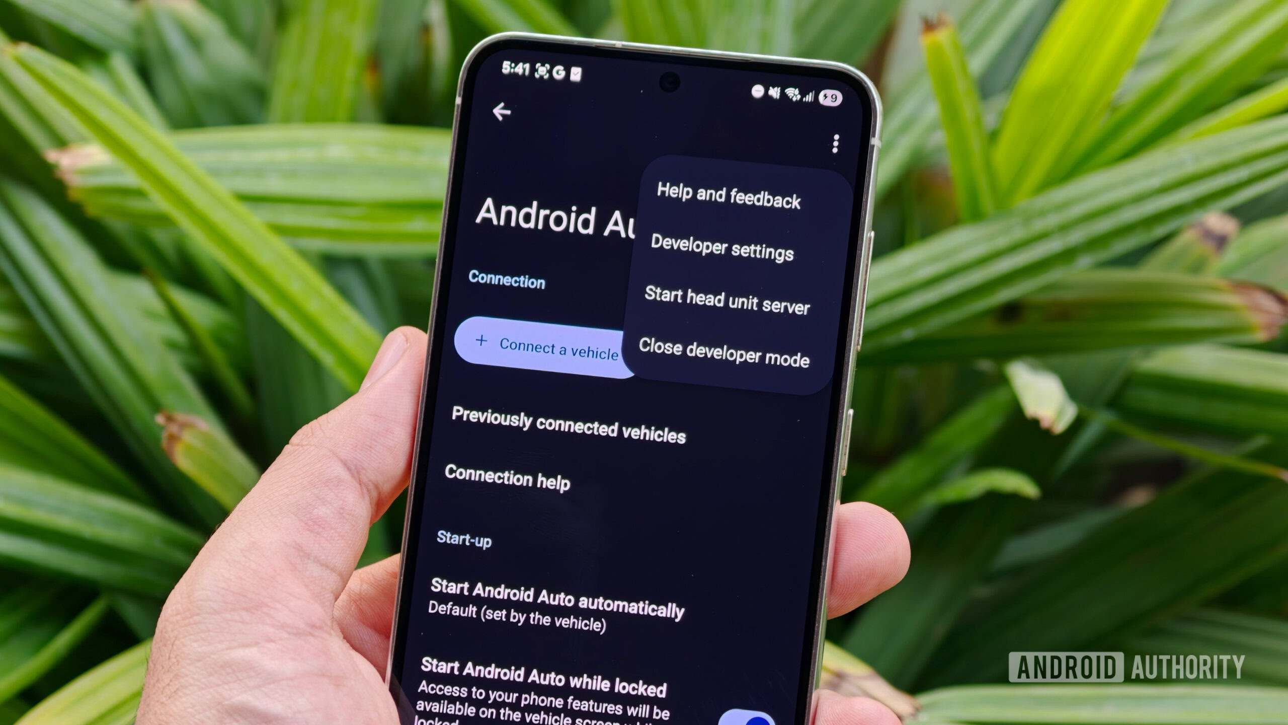 aplicativos de terceiros para Android Auto 1 Uma mão segurando um smartphone mostrando a tela de configurações do Android Auto com o menu flutuante de configurações do desenvolvedor aberto