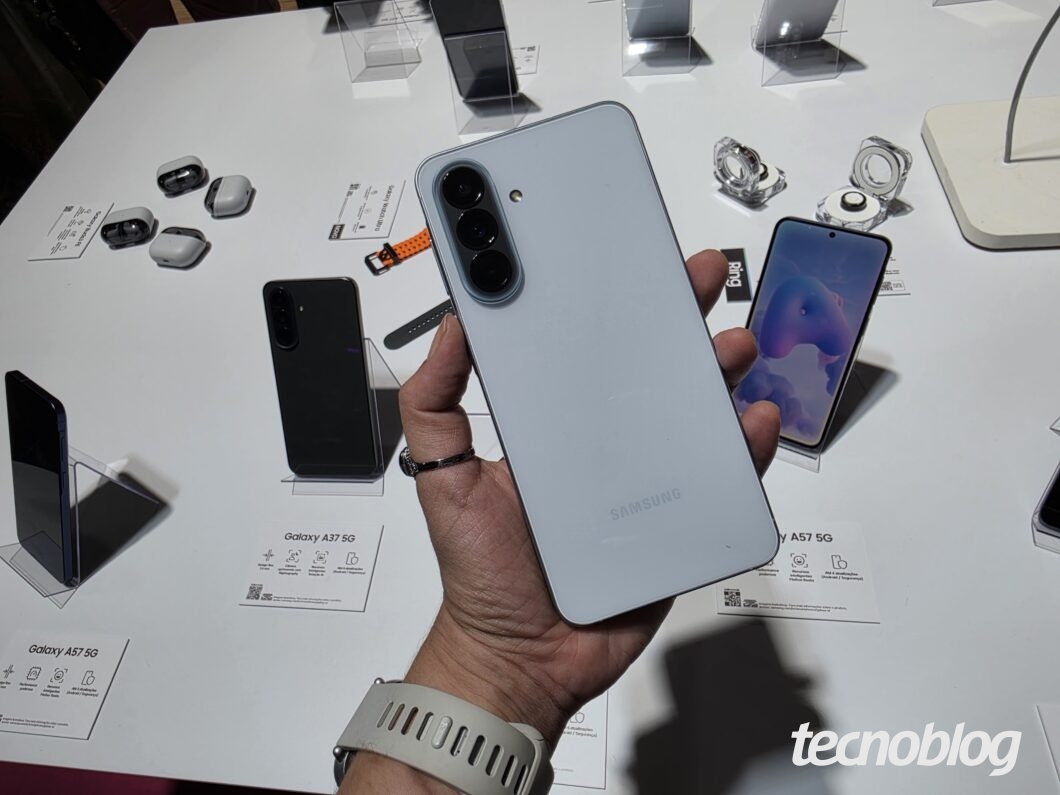 Mão exibindo um Samsung Galaxy A57 5G com câmera tripla em uma bancada com outros modelos e acessórios